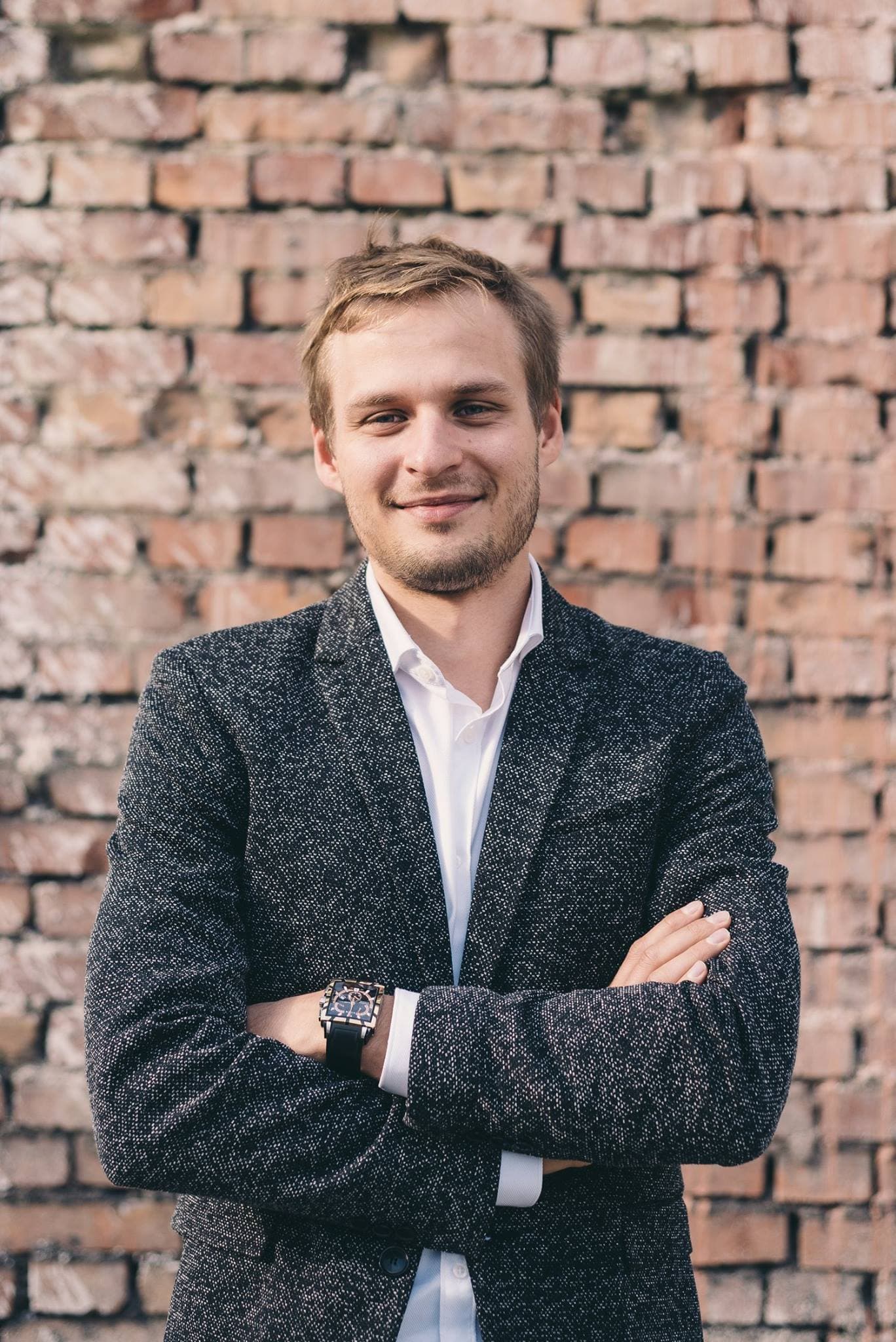 Smiling man with arms crossed wearing a textured blazer against a weathered brick wall.