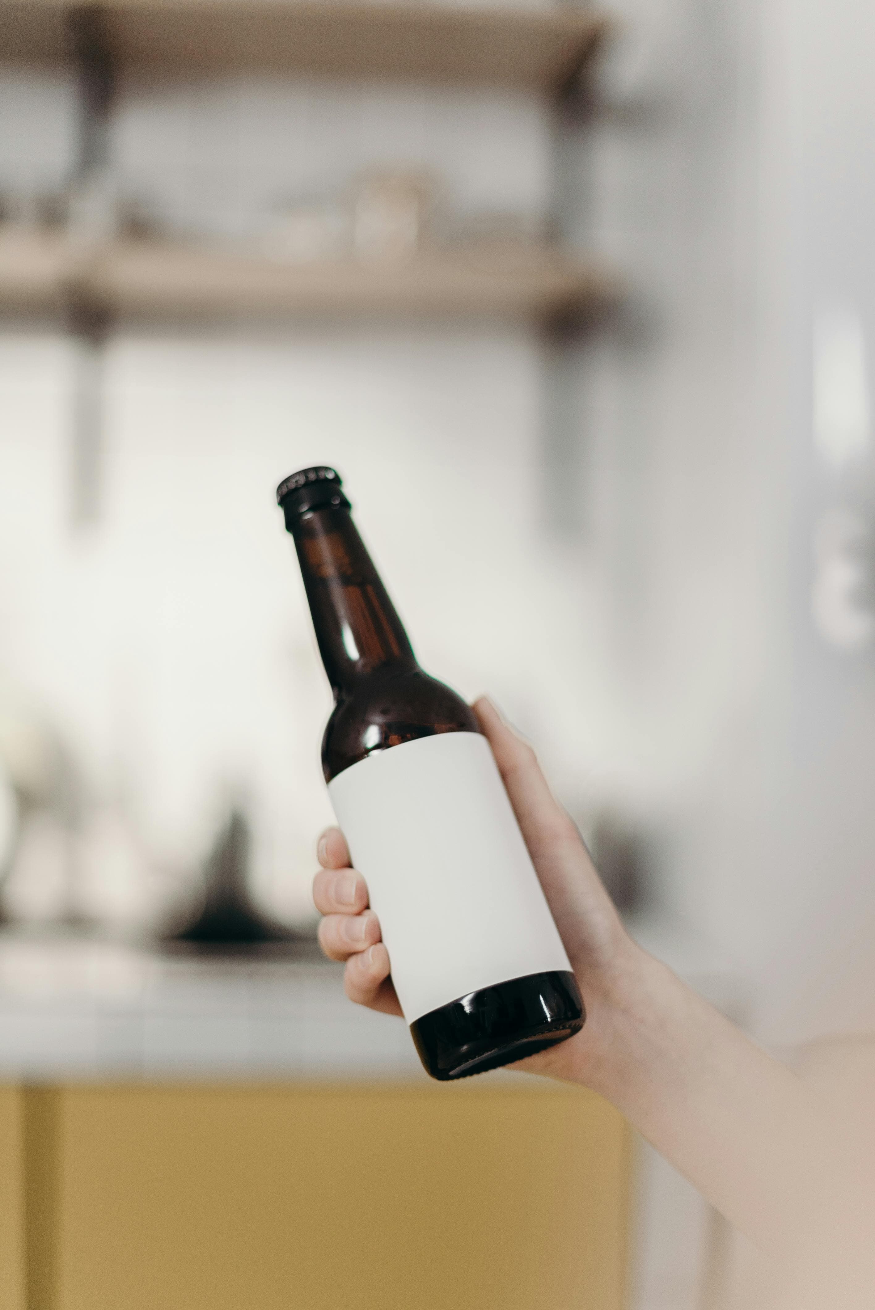 Hand holding a brown glass bottle with a blank white label in a blurred kitchen.
